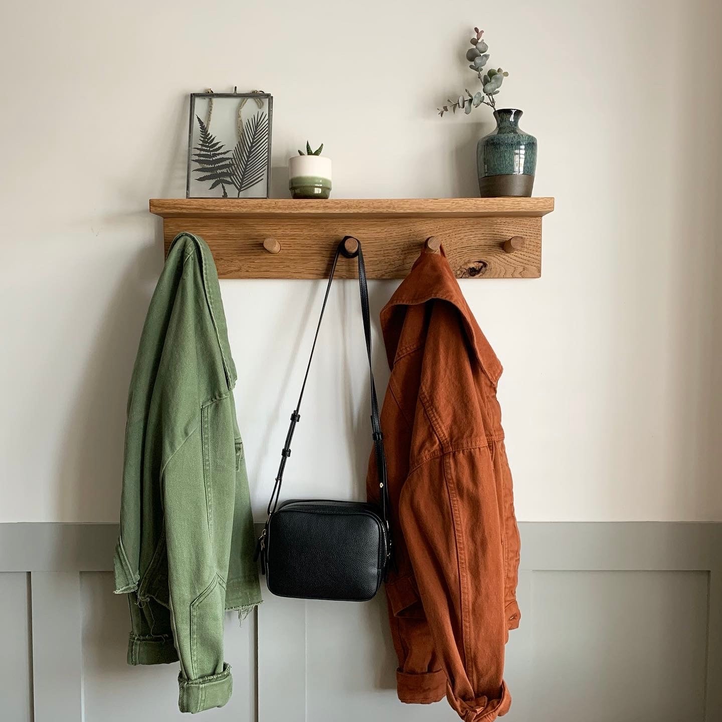 Wooden shelf with hooks holding a green jacket, black bag, and brown coat against a white wall.